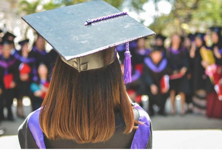 View of graduate's head from the back. They are wearing cap and gown, and other graduates similarly dressed in the background.