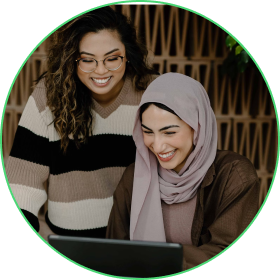 Two women, one wearing a headscarf, looking at a laptop, smiling.